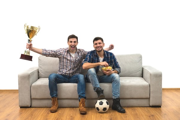 The two happy men with a cup watch a football on the sofa