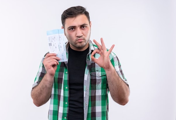 Young traveler man in checked shirt holding air tickets looking confident doing ok sign standing over white wall